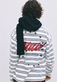 Person mit lockigem Haar trägt ein langärmliges, gestreiftes weißes Hemd mit Sternen und rotem Text auf dem Rücken, einen schwarzen Schal über die Schultern drapiert.