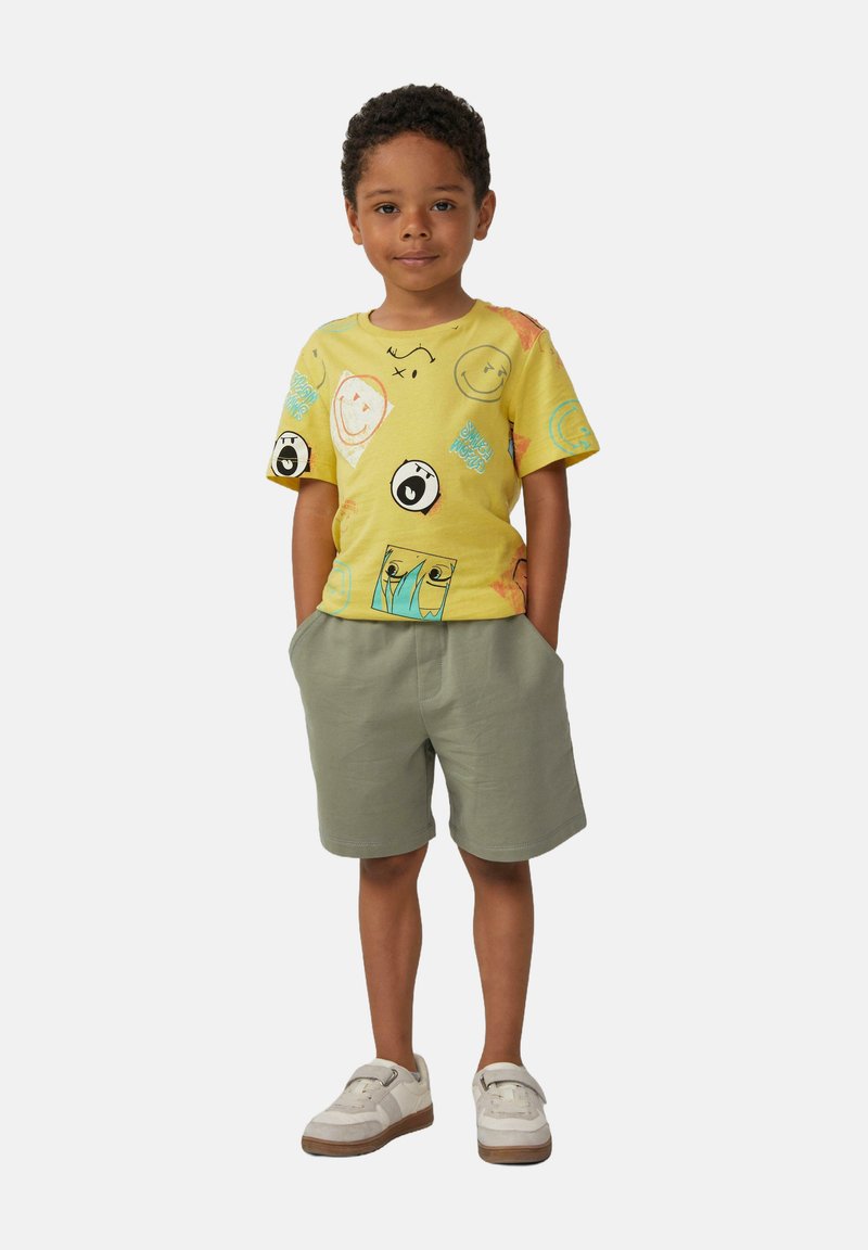 T-shirt di cotone giallo con stampe di volti giocosi, abbinato a pantaloni corti verdi. L'outfit presenta una vestibilità comoda e un tessuto confortevole.