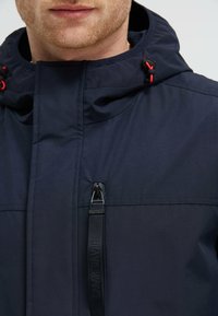 Granatowa kurtka z kieszenią na zamek błyskawiczny z przodu, regulowanym kapturem ze sznurkiem ściągającym i kontrastującymi czerwonymi akcentami. Gładka faktura i lekka konstrukcja.