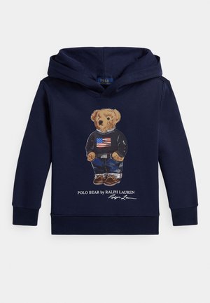 Felpa con cappuccio blu navy con grafica di un orso che indossa un maglione con la bandiera americana, jeans e scarpe marroni, con la scritta "Polo Bear by Ralph Lauren" sotto.