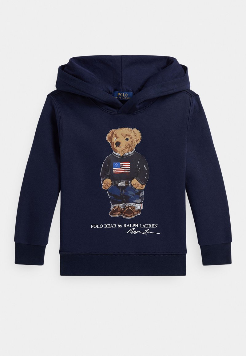 Tumši zils sporta krekls ar lāča attēlu, kas valkā džemperi ar Amerikas karogu, džinsus un brūnas kurpes, zemāk teksts "Polo Bear by Ralph Lauren".