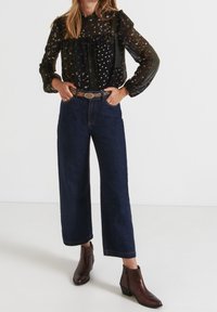 Schwarz gemusterte Bluse mit langen Puffärmeln, kombiniert mit dunkelblauen weit geschnittenen Jeans und braunen Leder-Ankle-Boots. Der Gürtel weist metallische Akzente auf.