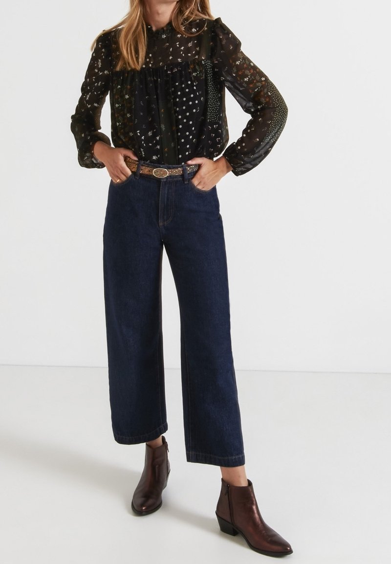 Schwarz gemusterte Bluse mit langen Puffärmeln, kombiniert mit dunkelblauen weit geschnittenen Jeans und braunen Leder-Ankle-Boots. Der Gürtel weist metallische Akzente auf.