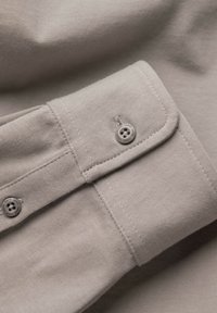 Gros plan sur un poignet de chemise beige avec un bouton fermé et des coutures détaillées sur un tissu lisse.