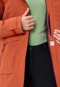 Giacca impermeabile arancione con zip, aperta per mostrare una maglietta verde sotto. Dettagli notevoli includono un accento rosso sul polsino e una tasca laterale.