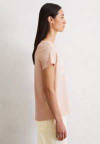 Korte mouwen, lichtroze katoenen T-shirt met een ronde halslijn en witte bedrukte tekst, gecombineerd met lichtgekleurde broek, zijaanzicht weergegeven.