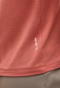 Κοντινή λήψη της υφασμάτινης, σκουρόχρωμης πορτοκαλί τελειώματος πουκάμισου με λευκό λογότυπο "DRYTON" πάνω από τη ραφή μπεζ παντελονιού.