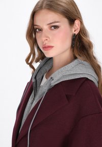 Cappotto in lana bordeaux con un hoodie grigio sotto, featuring un colletto a faccetta e chiusura con zip, tessuto testurizzato e dettagli argentati.