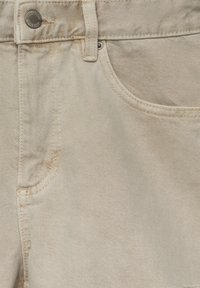 Gros plan sur un jean en denim beige montrant la ceinture, un bouton métallique portant l'inscription "Pull&Bear", une passoire pour ceinture, et une poche avant incurvée avec des coutures visibles.