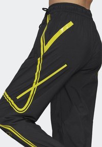 Černé sportovní kalhoty s žlutými abstraktními pruhy, se zipovou boční kapsou a pasem na šňůrku, nošené v pohybu.