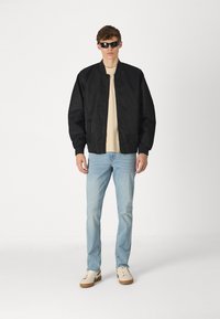 Black bomber jacket, lightweight material, ribbed cuffs, beige t-shirt, light blue jeans, and white sneakers with tan soles.