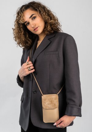 Sac bandoulière beige avec rabat en daim et corps en cuir, présentant un logo décoratif et une fine sangle, porté sur un blazer foncé.