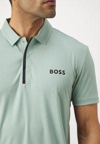Vihreä polo-paita, jossa on tekstuurikuvio, musta yksityiskohta nappilistassa ja "BOSS"-logo paksulla mustalla kirjaimella rintakehässä.