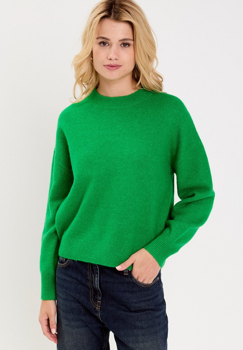 Groen gebreide trui met een ronde hals, licht afhangende schouders en geribbelde manchetten. Gecombineerd met donkere jeans, die een ontspannen pasvorm tonen.