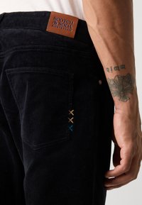 Black corduroy pants with a brown leather waistband label. Features decorative embroidered crosses in gold, blue, and brown on the back pocket.