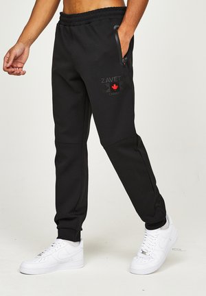 Sorte joggerbukser med lynlåslomme og rød ahornbladlogo på venstre lår, båret med hvide sneakers på lyse baggrund.