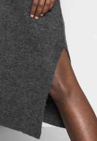 Abito grigio lavorato a maglia con uno spacco laterale, che mostra una consistenza morbida e una vestibilità rilassata su uno sfondo semplice. Tono di pelle uniforme visibile.