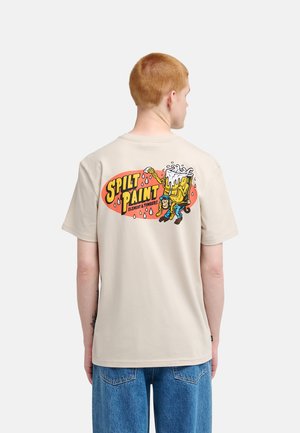 Młoda osoba z krótkimi czerwonymi włosami, nosząca beżowy t-shirt z kolorową grafiką z kreskówką "Spilt Paint" oraz niebieskie dżinsy, widziana od tyłu.