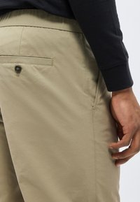 Κοντινό πλάνο ενός ατόμου που φοράει μπεζ παντελόνια με κουμπωμένη πίσω τσέπη, στέκεται με το δεξί του χέρι χαλαρό στο πλάι.