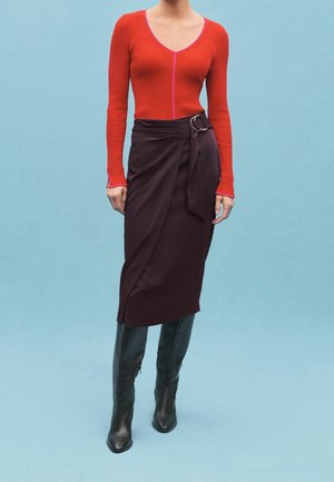 Haut rouge à manches longues côtelé avec une couture rose, associé à une jupe portefeuille bordeaux avec un nœud latéral et un accent anneau argenté ; bottes noires jusqu'aux genoux.