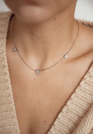 Zilveren ketting met drie bloemvormige hangers met witte bloemblaadjes. Gedragen op een neutraal gekleurde, gebreide trui met V-hals.