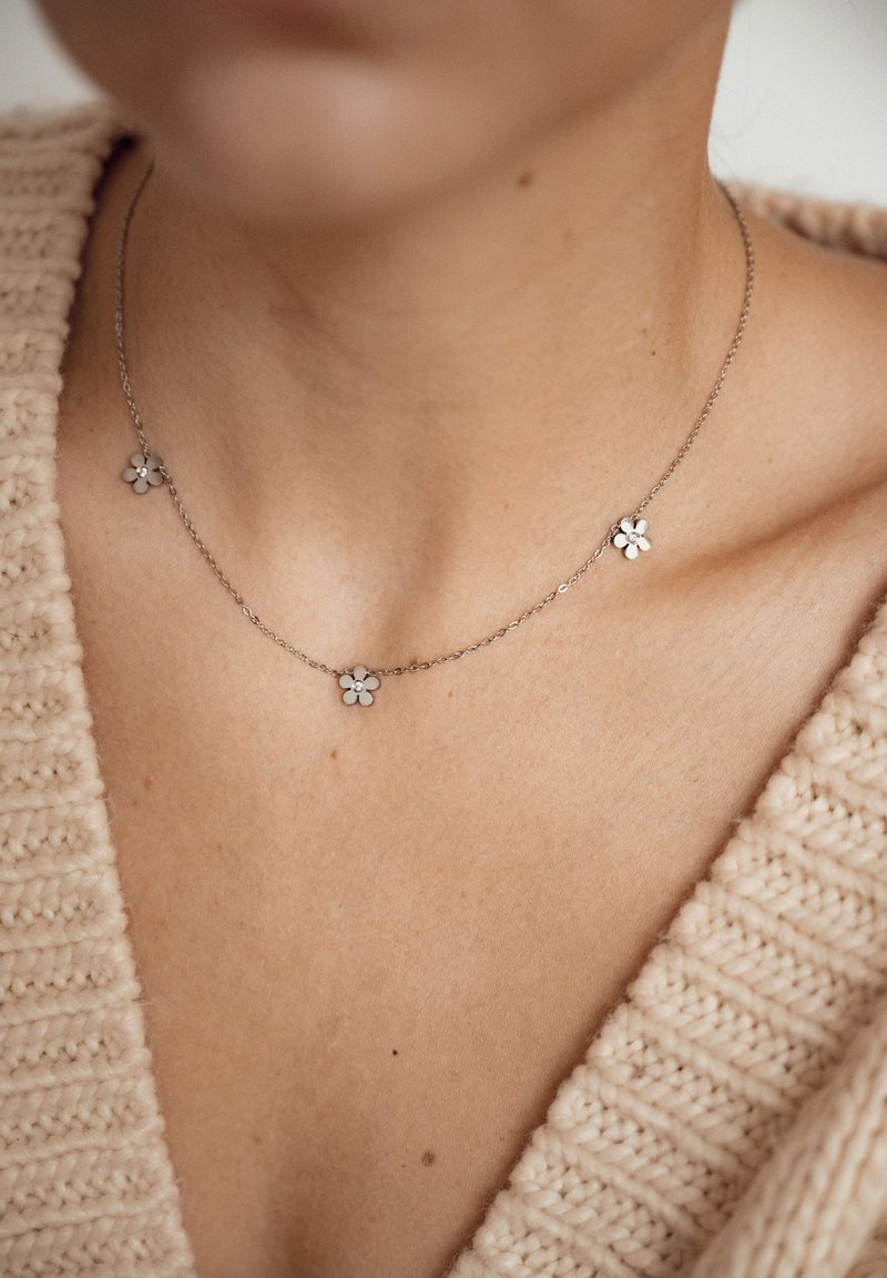 Collar de cadena de plata con tres colgantes florales con pétalos blancos. Llevado sobre un suéter de punto de color neutro con escote en V.