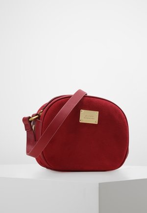 Kleine rode suède crossbodytas met een afgeronde vorm, verstelbare riem en een gouden merkplaatje aan de voorkant.