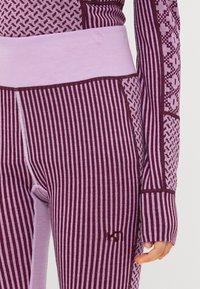 Mallas en púrpura y burdeos, con rayas verticales y patrones geométricos, un ancho cinturón rosa y detalles de costura contrastantes.
