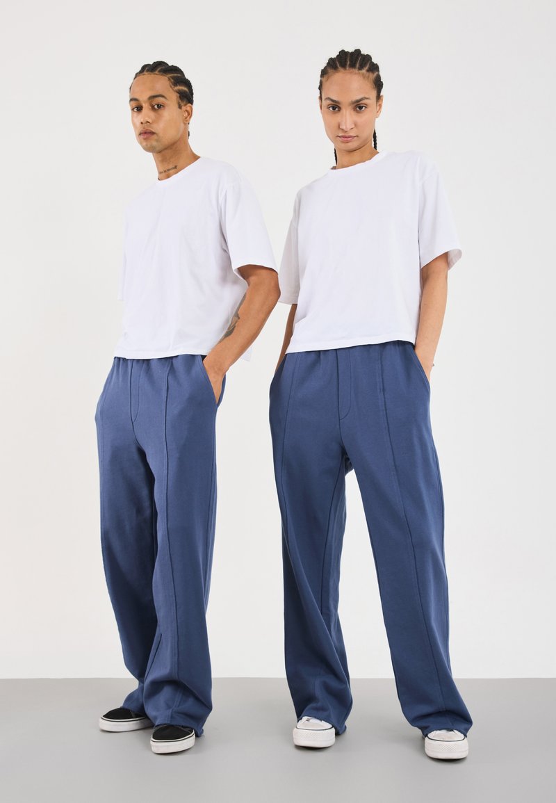 T-shirt bianchi oversize abbinati a pantaloni larghi blu, con tasche laterali e cuciture a contrasto, indossati con sneaker basse.