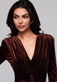 Sametové šarlatové šaty s hlubokým V výstřihem, dlouhými rukávy a nařasením v pase. Na modelce se objevuje jemný úsměv.