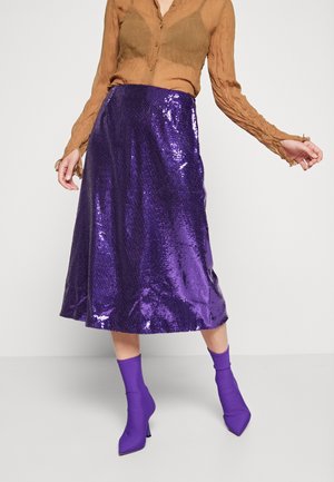 Kvinde iført en skinnende lilla paliet midi-nederdel, matchende lilla hælede sokkestøvler og en gennemskinnelig brun bluse med lange ærmer.