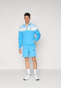 Uma jaqueta azul clara com fecho éclair, com riscas brancas e logo, emparelhada com calções correspondentes. O tecido parece suave; ambas as peças apresentam bolsos laterais.