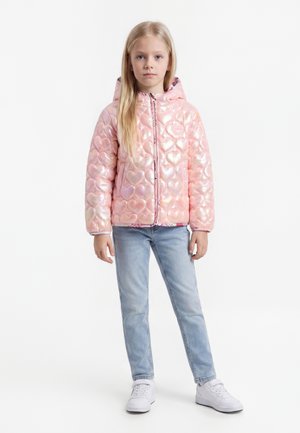 Ein junges Mädchen steht und trägt eine glänzende rosa Steppjacke mit Herzmuster, hellblaue Jeans und weiße Sneakers vor weißem Hintergrund.