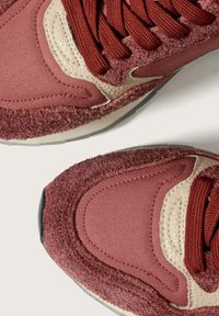 Närbild av bordeauxfärgade sneakers med texturerad mocka, tygpaneler, beige läderdetaljer och matchande bordeauxsnören på en ljus yta.