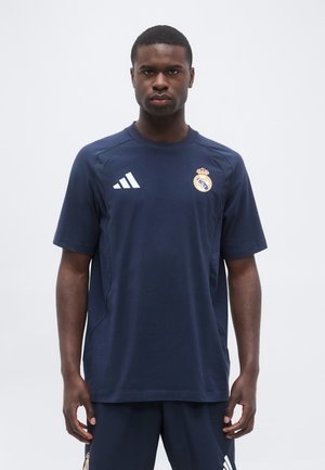 Homme debout face à nous portant un t-shirt et un short bleu marine Adidas avec les logos du club de football du Real Madrid sur la poitrine et la cuisse.