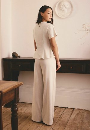 Frau in beigen, lockeren Hosen und kurzärmeligem Oberteil steht drinnen neben einem dunklen Holzkonsoltisch mit dekorativer runder Wandskulptur.