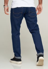 Jeans de mezclilla azul oscuro con corte recto, que cuentan con dos bolsillos traseros con costura amarilla y puños doblados sobre zapatillas negras.