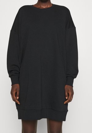 Osoba nosząca luźną, czarną sukienkę w formie bluzy z długimi rękawami, z prążkowanymi mankietami i dołem, stojąca na jednolitym tle.