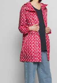 Czerwona wodoodporna kurtka z powtarzającym się wzorem psów, z kapturem, zapięciem na zamek i elastycznymi mankietami. Lekki materiał o gładkiej teksturze.