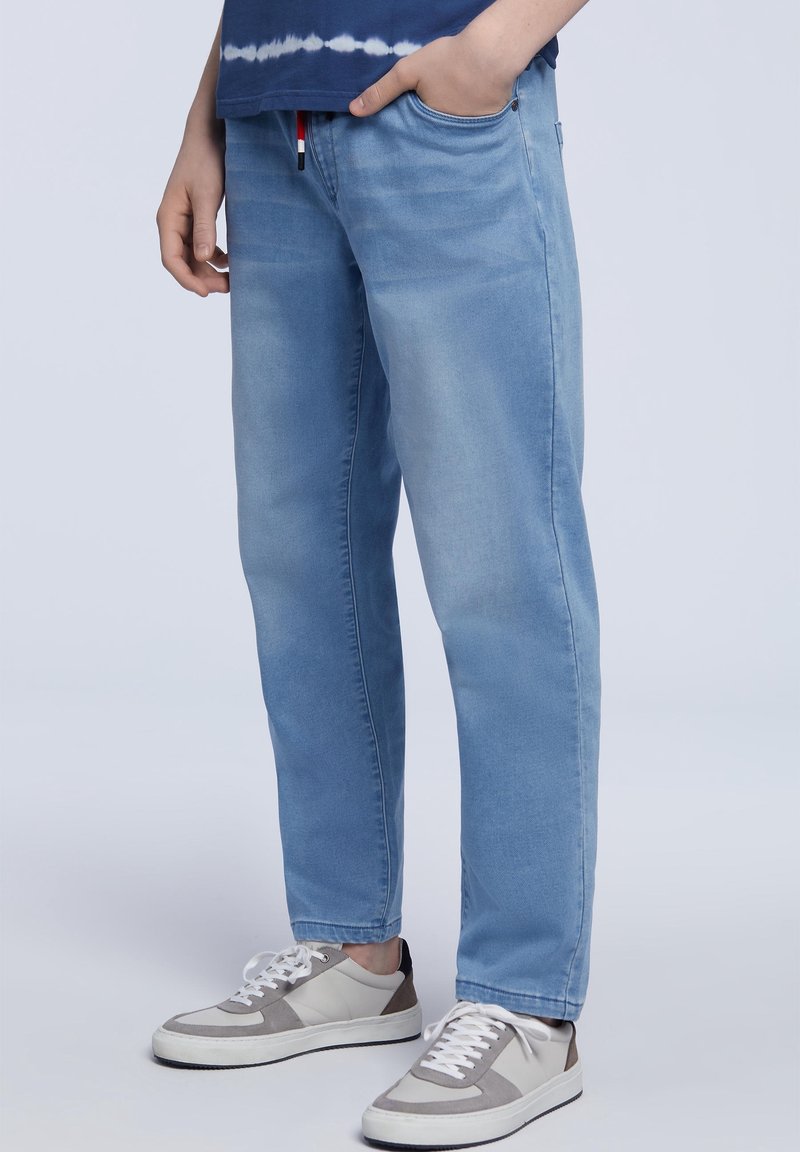 Personne portant un jean en denim bleu clair et des baskets blanches avec des accents gris, debout, une main dans la poche, sur un fond uni.