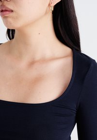 Haut ajusté bleu marine avec un décolleté large et des manches longues. Texture de tissu lisse, avec des détails de couture subtils le long du décolleté.