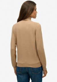 Donna con lunghi capelli castani che indossa un maglione a collo alto beige e jeans blu, di spalle su uno sfondo bianco uniforme.