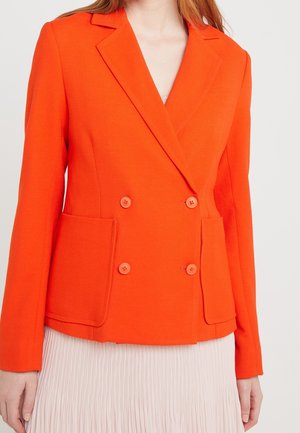 Kvinde iført en livlig orange dobbeltradet blazer med store forlommer over et lyst plisseret skørt.