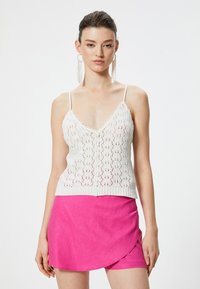Top de punto blanco sin mangas con un diseño en forma de V, que presenta un patrón de calado y cierre de botones. Combinado con una falda envolvente de color rosa brillante.