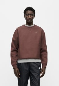 Μπλούζα φούτερ σε βυσσινί χρώμα με στρογγυλή λαιμόκοψη και ελαστικά μανίκια, διακοσμημένη με μικρό λευκό λογότυπο Nike. Συνδυάζεται με μαύρο αθλητικό παντελόνι.