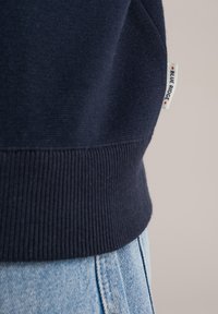 Mornarsko plavi pulover s rebrastim rubom i manžetama, s tkanom oznakom "Blue Ridge" sa strane. Zoom prikaz koji pokazuje teksturu i detalje dizajna.