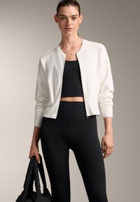Bela bomberska jakna s prilagojenim dizajnom, rebrastimi manšetami in zadrgo spredaj. Oblečena nad črno majico brez rokavov in hlačami z visokim pasom. Črna torba, ki jo držite v roki.