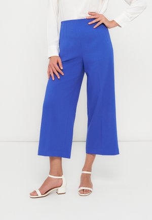 Femme portant un pantalon large court bleu vif, un chemisier blanc à manches longues, et des sandales blanches à talons bloc, debout avec une main sur la hanche.