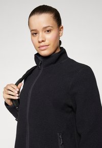 Black fleece jacket with a high collar, zip closure, and textured surface. Features side pocket and is worn with a shoulder strap.