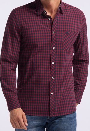 Mężczyzna w koszuli na guziki z długim rękawem o czerwono-czarnej kratce i kieszenią na piersi, w połączeniu z niebieskimi dżinsami.